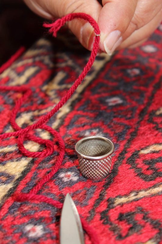 Hand repairing a traditional Persian rug with red thread and a small metal tool.