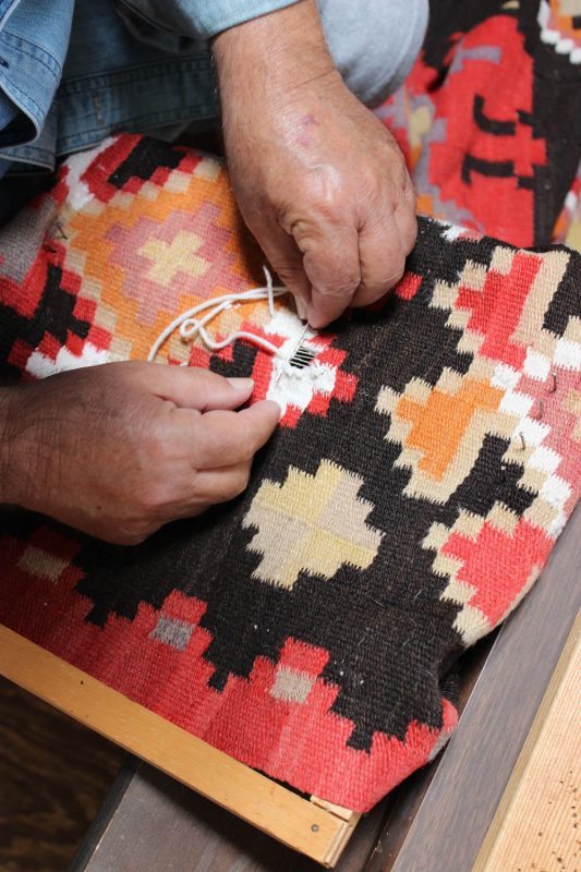Expert hand repairing a vibrant, patterned rug with needle and thread in a workshop.