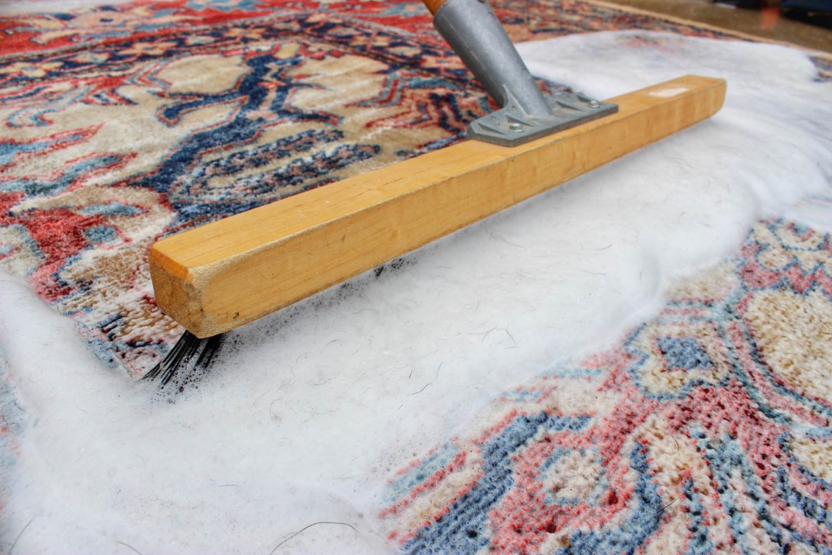 A wooden-handled brush spreading white cleaning foam across a red and blue patterned Oriental rug during a hand wash.