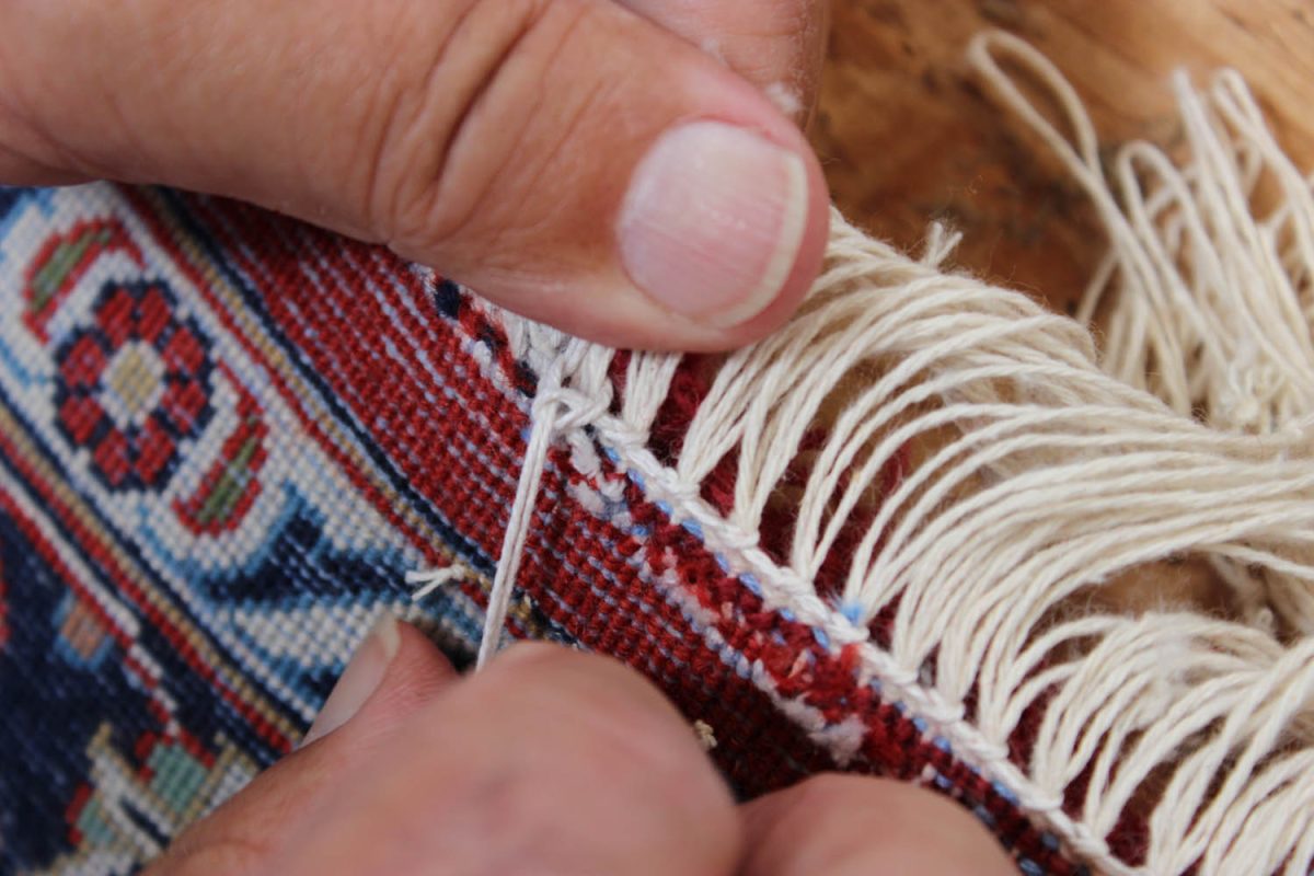Hands knotting new fringe onto the edge of a red and blue rug